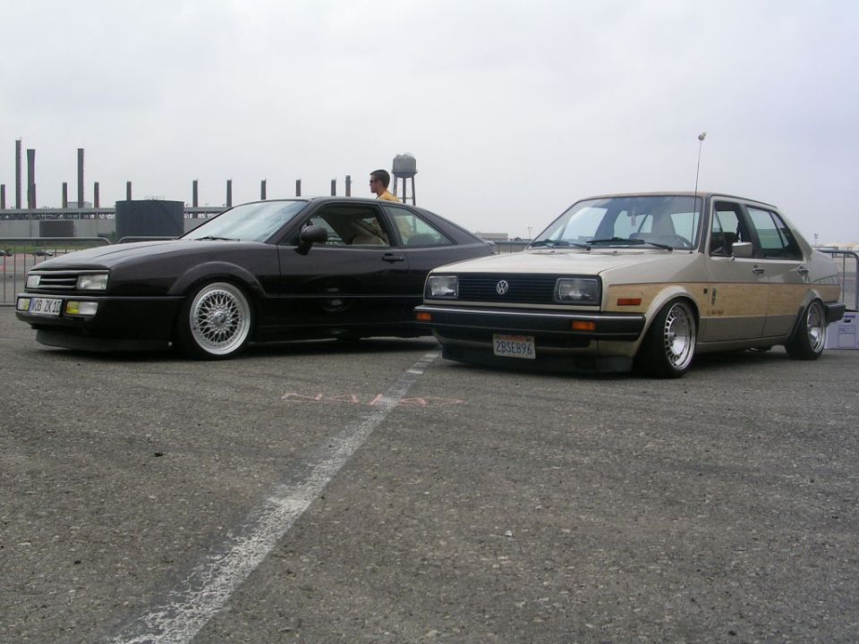 Two lowered Volkswagen cars, a dark coupe and a tan sedan with wood paneling, parked on asphalt
