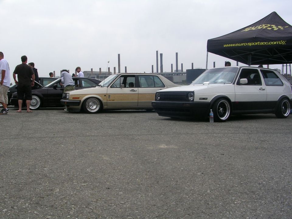 Three cars parked on asphalt, two Volkswagen Golfs and a sedan with wood paneling. A tent is set up behind the white Golf