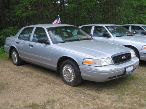 1991 Ford LTD Crown Victoria - A silver sedan with an American flag on its roof is parked next to other similar vehicles.