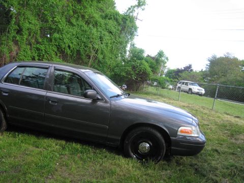1991 Ford LTD Crown Victoria - A dark sedan is parked on a grassy incline with a white SUV visible in the distance behind a chain-link fence.