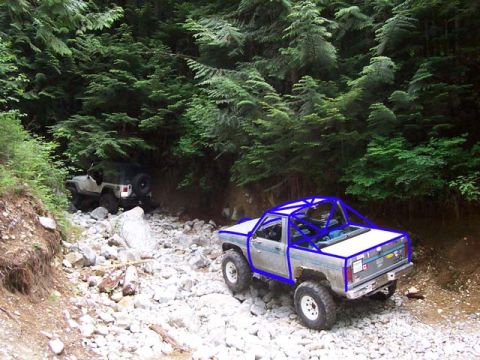 1984 Ford Bronco II - Two off-road vehicles navigate a rocky path through a dense forest.