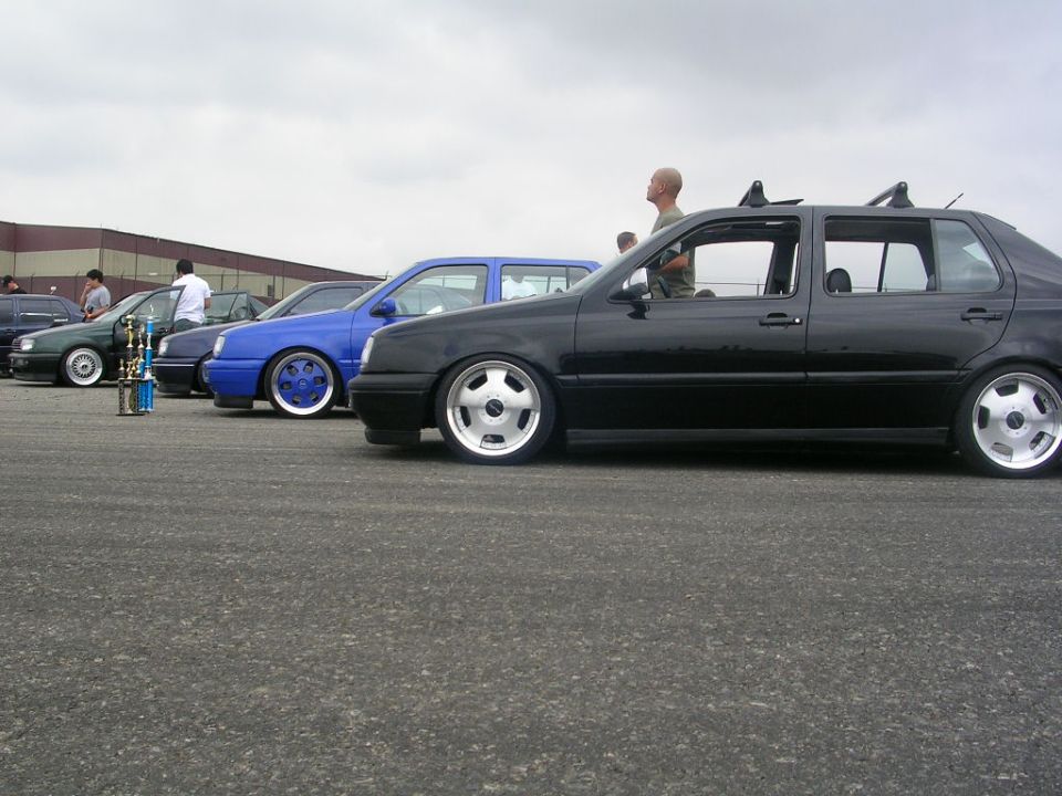 Several lowered sedans parked in a row, one black, one blue, one green. Trophies are visible in front of the green car