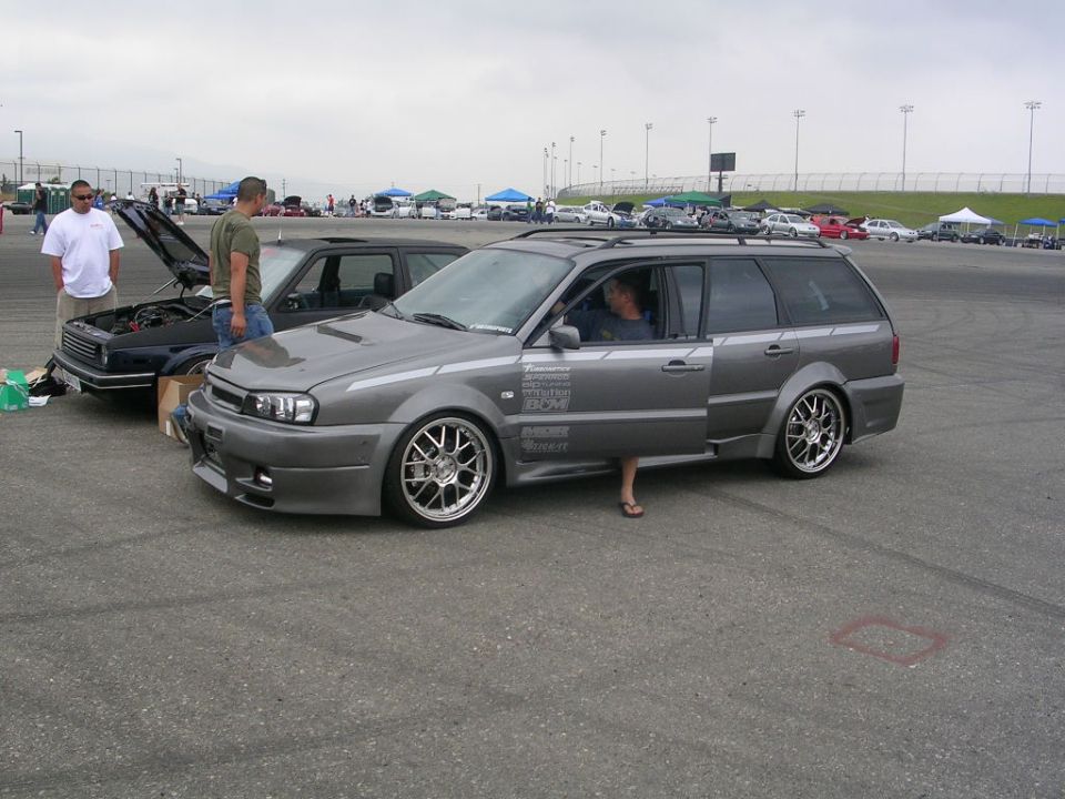 Modified gray station wagon with racing decals on the door, parked next to a black car with hood open. Two men stand nearby