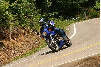 1985 Ford Crown Victoria LTD - A motorcycle with two riders navigates a sharp turn on a scenic, wooded highway.