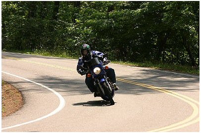 1985 Ford Crown Victoria LTD - A motorcyclist navigates a sharp turn on a scenic road, surrounded by lush green trees.