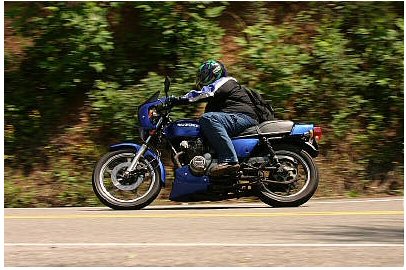 1985 Ford Crown Victoria LTD - A rider on a blue motorcycle travels down a road with trees lining the side.
