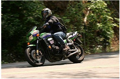 1985 Ford Crown Victoria LTD - Rider on a motorcycle navigates a paved road surrounded by trees.