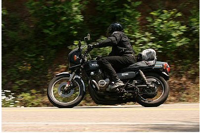 1985 Ford Crown Victoria LTD - A rider on a black Suzuki motorcycle cruises down a tree-lined road.