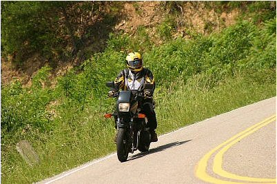 1985 Ford Crown Victoria LTD - A motorcycle rider navigates a curve on a scenic road, surrounded by lush greenery.