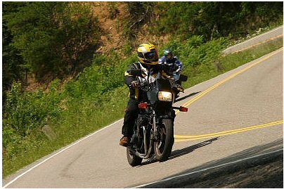 1985 Ford Crown Victoria LTD - Motorcyclists navigate a scenic, winding road, with one rider in the foreground and another following behind.
