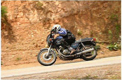 1985 Ford Crown Victoria LTD - A motorcyclist navigates a winding road, leaning into a turn with a rocky hillside to their left.