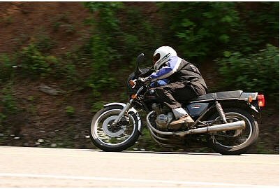 1985 Ford Crown Victoria LTD - A motorcycle rider navigates a road, with a blurred background of trees and foliage.