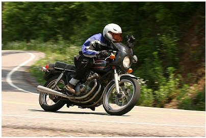 1985 Ford Crown Victoria LTD - A rider navigates a curve on a motorcycle, with trees blurring in the background.