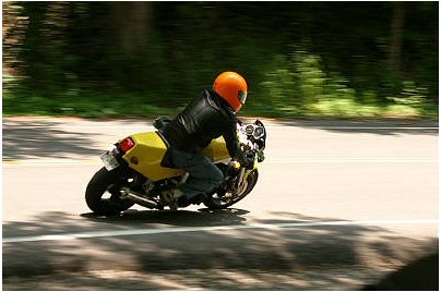 1985 Ford Crown Victoria LTD - A motorcycle rider in an orange helmet and black jacket speeds down a tree-lined road.