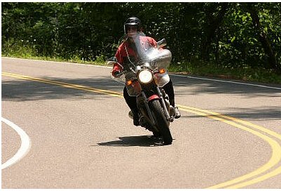 1985 Ford LTD Crown Victoria - A rider on a motorcycle navigates a sharp turn on a scenic road, with trees lining the asphalt.