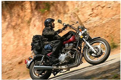 1985 Ford LTD Crown Victoria - A rider on a motorcycle navigates a winding road with a dirt embankment on the side.