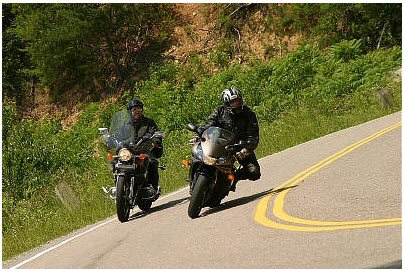 1985 Ford LTD Crown Victoria - Two motorcyclists navigate a curve on a scenic road, with lush greenery lining the roadside.