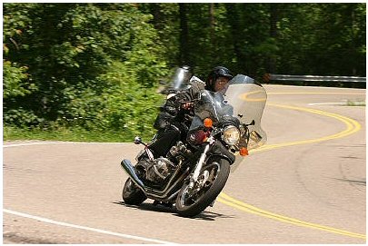 1985 Ford LTD Crown Victoria - A motorcycle navigates a sharp turn on a scenic road surrounded by trees.