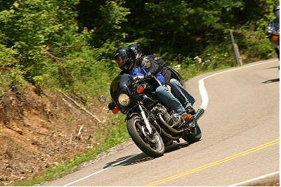1985 Ford LTD Crown Victoria - Two people ride a motorcycle on a winding road through a wooded area.