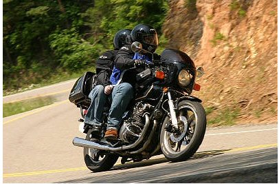 1985 Ford LTD Crown Victoria - Two individuals ride a black motorcycle along a scenic, winding road, with a rocky embankment to their right.