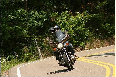 1985 Ford LTD Crown Victoria - Two people ride a motorcycle on a curved road surrounded by trees.
