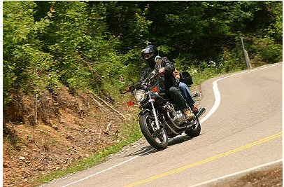 1985 Ford LTD Crown Victoria - Two people ride a motorcycle on a scenic, winding road surrounded by trees.