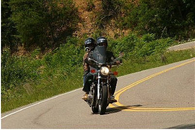 1985 Ford LTD Crown Victoria - A motorcycle carries two riders along a scenic, curving road bordered by lush greenery.