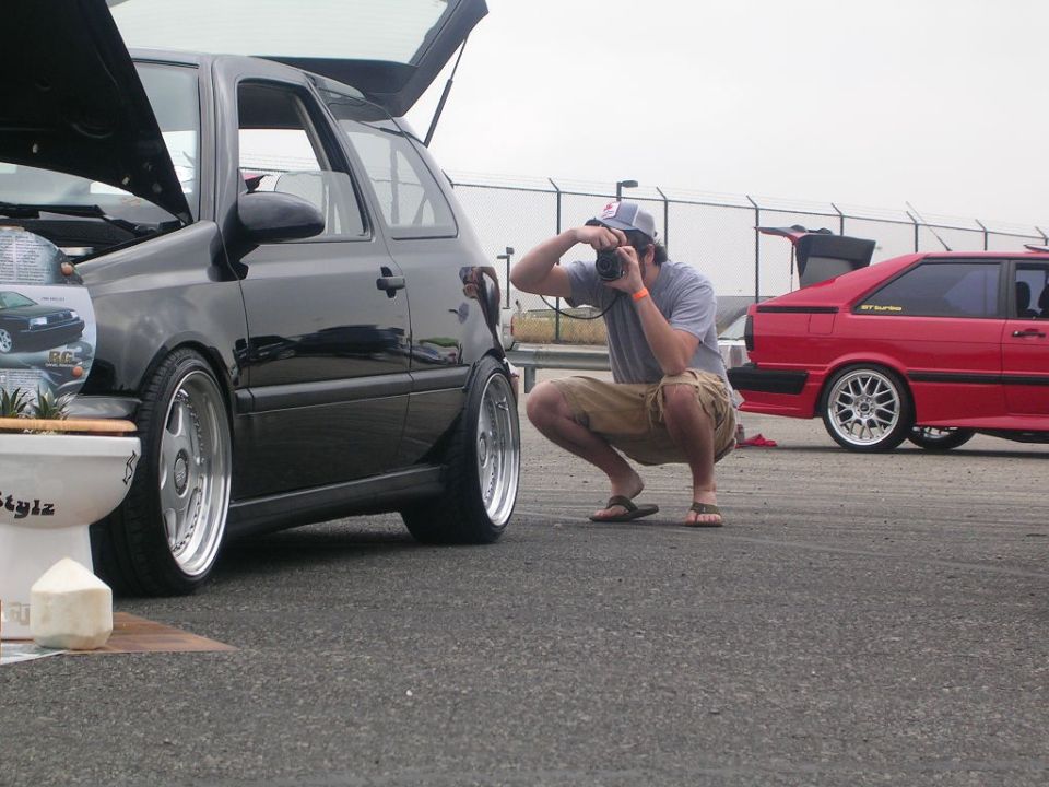 Black Volkswagen Golf GTI with hood and trunk open, photographer crouching nearby. Red Audi coupe in background