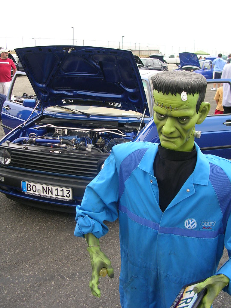 Frankenstein mannequin in blue mechanic's jumpsuit next to a blue Volkswagen Golf with hood open. License plate reads BO NN 113