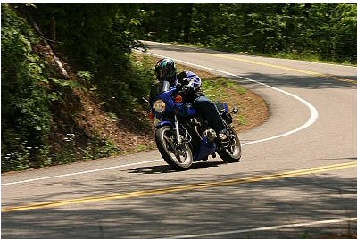 1985 Ford LTD Crown Victoria - A motorcyclist leans into a turn on a scenic, wooded highway.