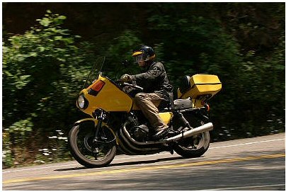 1985 Ford LTD Crown Victoria - A rider on a yellow motorcycle navigates a winding road surrounded by lush green trees.