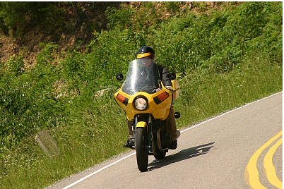 1985 Ford LTD Crown Victoria - A rider on a yellow motorcycle navigates a scenic road bordered by lush greenery.
