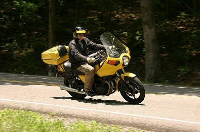 1985 Ford LTD Crown Victoria - A rider on a yellow motorcycle travels down a tree-lined road.