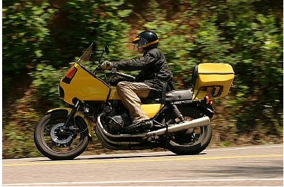 1985 Ford LTD Crown Victoria - A rider on a yellow motorcycle with a storage box travels down a scenic, tree-lined road.