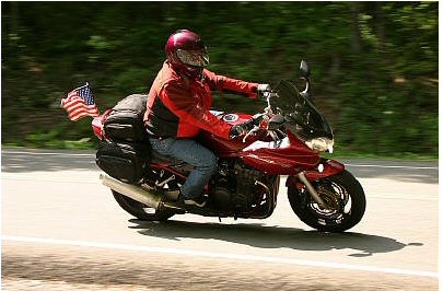1985 Ford LTD Crown Victoria - A rider on a red motorcycle, equipped with saddlebags and an American flag, cruises down a road surrounded by trees.