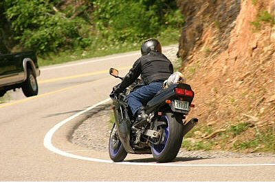 1985 Ford LTD Crown Victoria - A motorcycle rider navigates a curve on a scenic road with another vehicle visible.