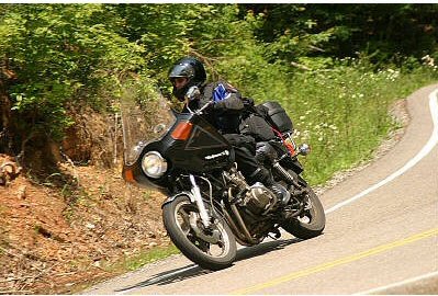 1985 Ford LTD Crown Victoria - A motorcyclist navigates a curve on a scenic road surrounded by lush greenery.
