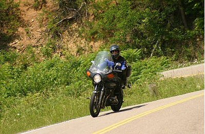 1985 Ford LTD Crown Victoria - A lone motorcyclist navigates a winding road surrounded by lush green foliage and a steep embankment.
