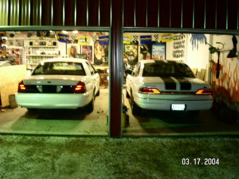 2001 Ford Crown Victoria - Two white Ford Crown Victorias are parked side-by-side in a garage, one with racing stripes.