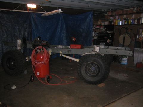 1978 Ford Bronco - Bare rolling chassis mid-restoration in a home garage: freshly painted gray frame rails on axles with mud-terrain tires and black steel whe…
