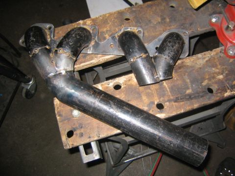 1978 Ford Bronco - A set of custom-fabricated tubular exhaust headers laid on a workbench, showing welded primary tubes and mounting flanges joining into a me…