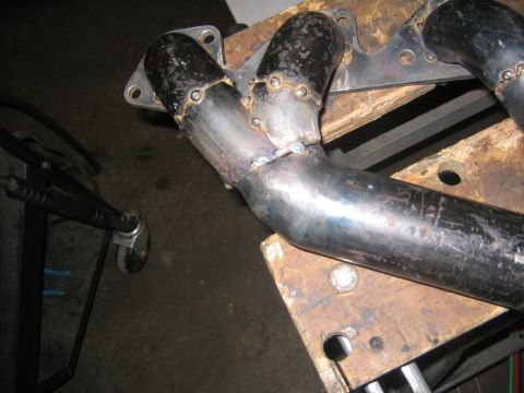 1978 Ford Bronco - A shop-fabricated tubular exhaust header lying on a scarred wooden workbench, with its primary tubes tack- and seam-welded into a collector…