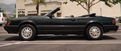 1985 Ford Mustang - This black convertible Mustang is parked on a street with a building and trees in the background.