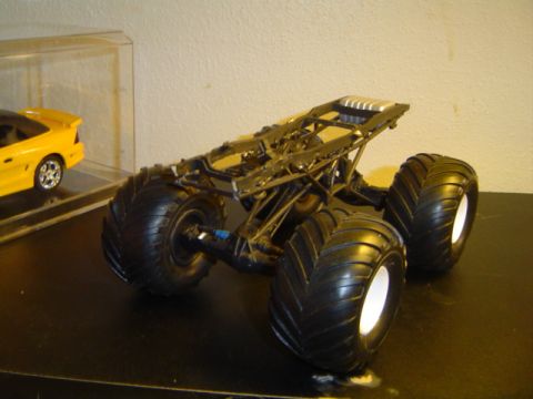1979 Ford Bronco - A model monster truck chassis sits on a black surface, with a yellow toy car in a display case in the background.