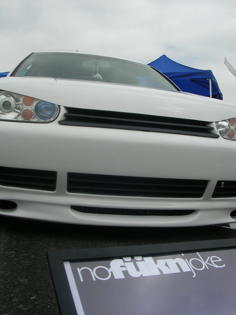 White Volkswagen Golf Mk4 front bumper with black grille and headlights. Sign in foreground reads 'nofüknjoke'