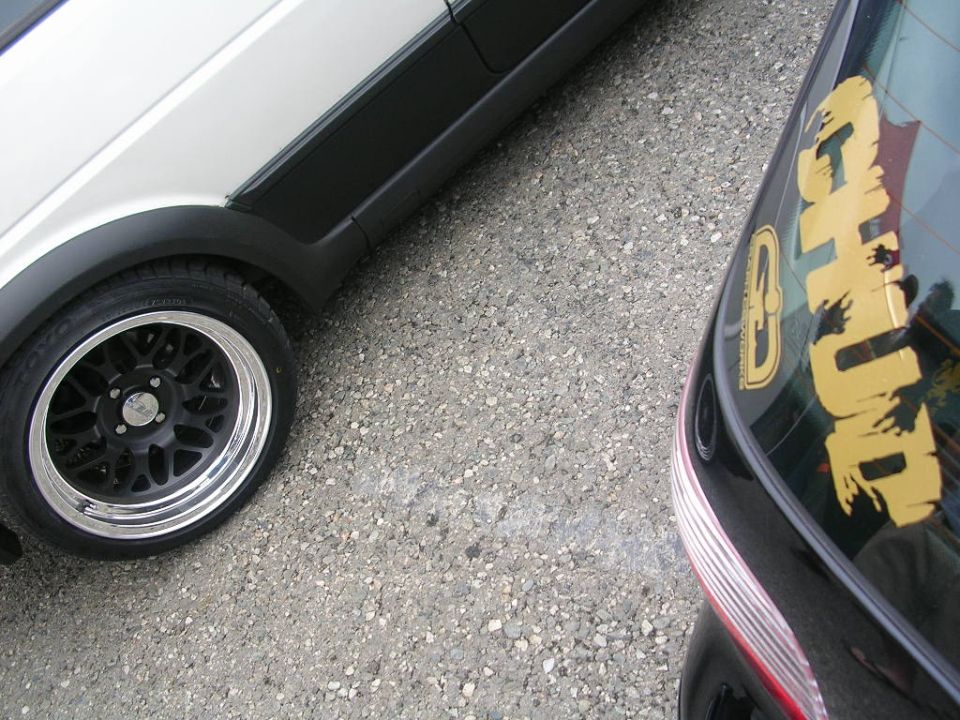 White vehicle fender and wheel with black trim, next to a black vehicle's rear with a large yellow sticker