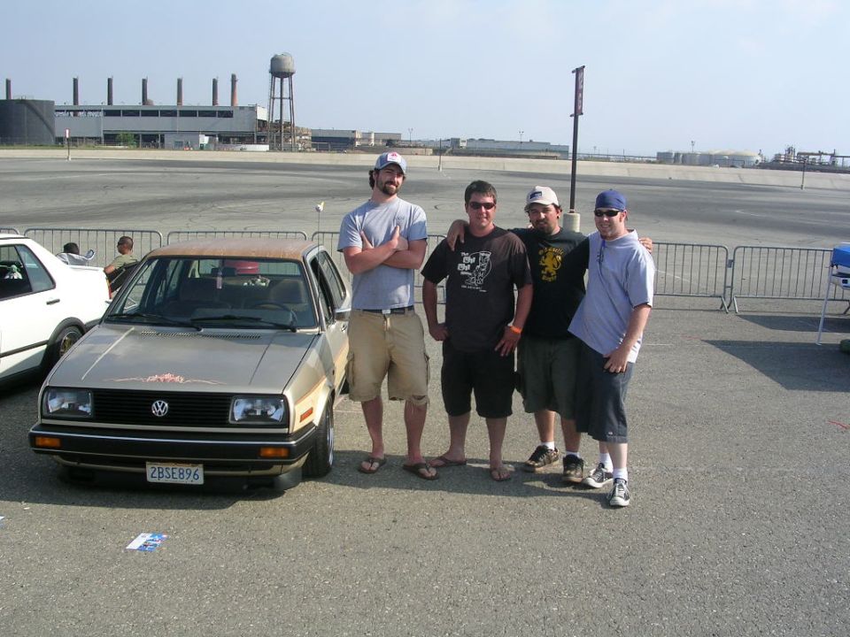 Lowered gold Mk2 VW Jetta with patina roof at an outdoor car meet, four men beside it