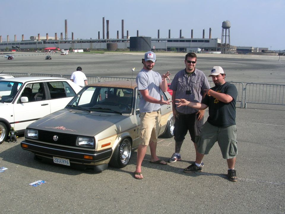 Three men posing next to a lowered tan Volkswagen Jetta sedan at an industrial lot