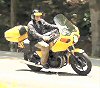 1985 Ford LTD Crown Victoria - A person rides a yellow motorcycle on a paved road, surrounded by trees.