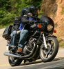 1985 Ford LTD Crown Victoria - Two individuals ride a black motorcycle along a scenic, winding road, with lush greenery visible on the side.